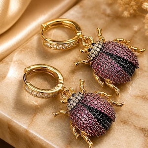 May include: A pair of gold hoop earrings featuring beetle-shaped charms. The hoops are encrusted with clear stones. The beetle charms are detailed with pink and black stones covering the wings and body, with gold-toned legs and head accents.