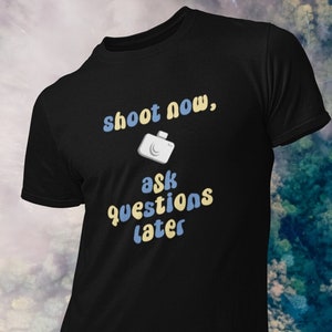 May include: Black t-shirt with the text "shoot now, ask questions later" in yellow and blue lettering. A camera icon is above the text.