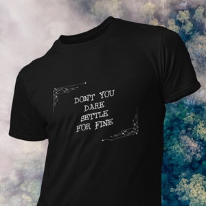 May include: Black t-shirt with the text "DON'T YOU DARE SETTLE FOR FINE" in white. The text is framed by decorative corner elements. The shirt is displayed against a blurred background of a forest.