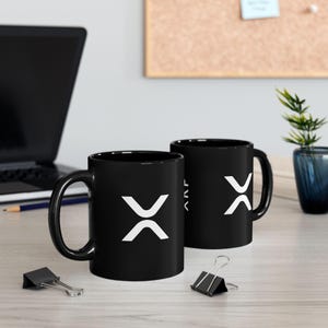 Puede incluir: Dos tazas de cerámica negra con el logotipo blanco de XRP se muestran sobre una superficie de madera clara. Un portátil, una planta y material de oficina están en el fondo. Las tazas tienen un acabado brillante.