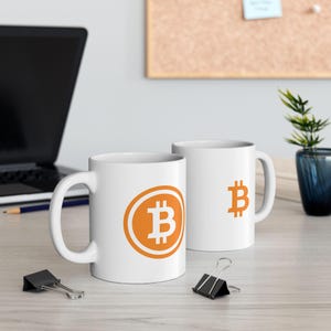 May include: Two white ceramic coffee mugs featuring the Bitcoin symbol in orange. One mug has the symbol within a circle, the other has the symbol on its own. Both mugs have a curved handle.