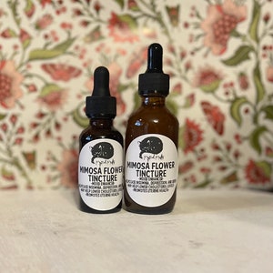 May include: Two amber glass bottles of Mimosa Flower Tincture with black droppers. Each bottle has a white label with a black cat illustration and text. The text includes "Mood Enhancer" and "Promotes Uterine Health".