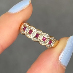 May include: A gold ring featuring a repeating circular pattern, with each circle containing a square-cut red ruby at the centre, surrounded by small, clear, round-cut white stones set into the metal band along the entire outer surface.