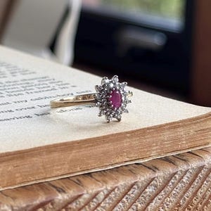 May include: A gold ring featuring a central oval-shaped red gemstone surrounded by a halo of small, clear, round-cut stones arranged in a starburst pattern. The ring is resting on the open page of a book with visible printed text and page number 107.
