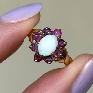 May include: A gold ring with an oval opal at its centre, encircled by smaller, round, deep pink gemstones. The ring's design is floral, with the gemstones forming a flower-like pattern. The ring is held between fingers.