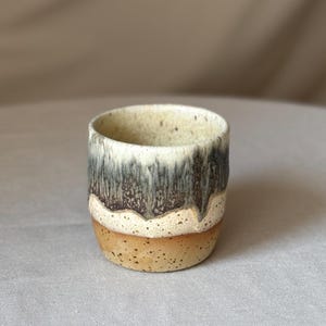 Op de afbeelding: Een kleine, handgemaakte keramische beker met een uniek, gelaagd ontwerp. De basis is een warm, gespikkeld bruin, overgaand in een crèmekleurige witte band, en afgewerkt met een donkere, gedruppelde glazuur. De beker is ongeveer 7.6 cm hoog.