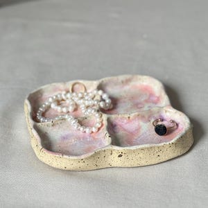 Könnte beinhalten: Ein handgefertigtes Schmucktablett aus Keramik mit drei Fächern. Das Tablett hat eine gesprenkelte beige Außenseite und eine rosa-weiße Innenseite. Eine Perlenkette und zwei Ringe sind auf dem Tablett ausgestellt.