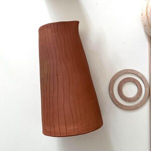 Pode incluir: Um jarro de cerâmica cor de terracota com um design texturizado de linhas verticais. O jarro tem uma forma curva e um pequeno bico. Dois porta-copos de madeira e uma pequena planta em vaso também são visíveis.