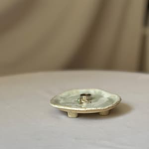 Op de afbeelding: Een kleine, gebroken witte keramische ringschaal met drie korte pootjes. De schaal heeft een enigszins onregelmatige vorm en een lichtgroene glazuur. Twee ringen liggen op de schaal. De schaal heeft een diameter van ongeveer 10 cm.