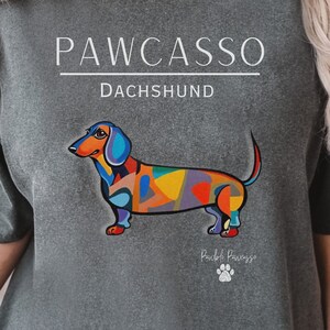 May include: A gray t-shirt with the text "PAWCASSO" and "DACHSHUND" printed on it. A colorful abstract dachshund is printed on the shirt. The text "Pawcasso" is printed below the dachshund.