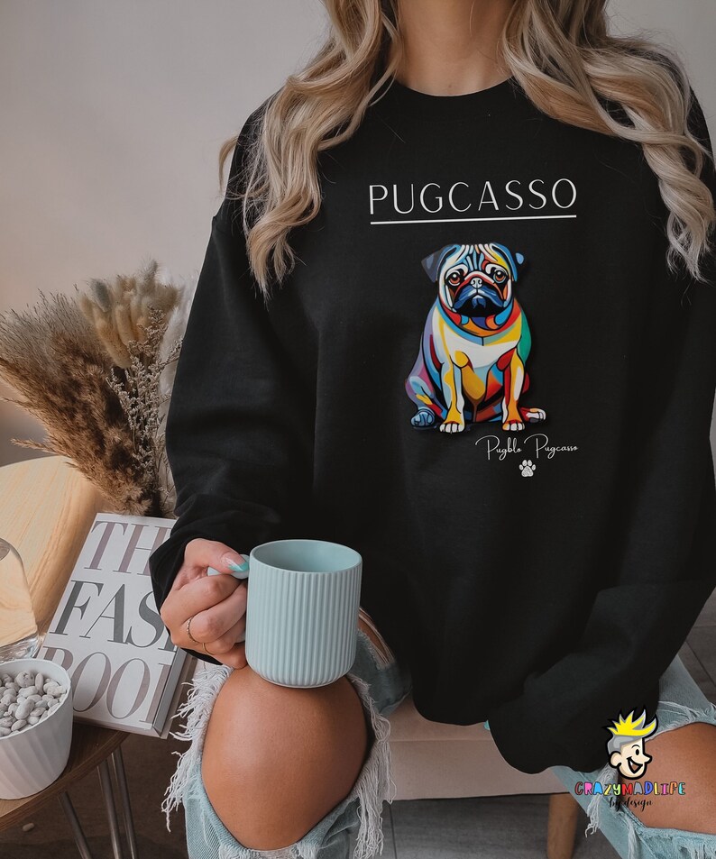 Puede incluir: Sudadera negra con una ilustraci&oacute;n de un carlino colorido y el texto "PUGCASSO" y "Puggle Picasso" en la parte delantera.