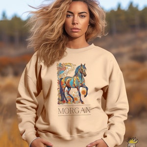 以下が含まれることがあります： ベージュのトレーナーに、カラフルな馬のグラフィックと、その下に「MORGAN」という名前がプリントされています。