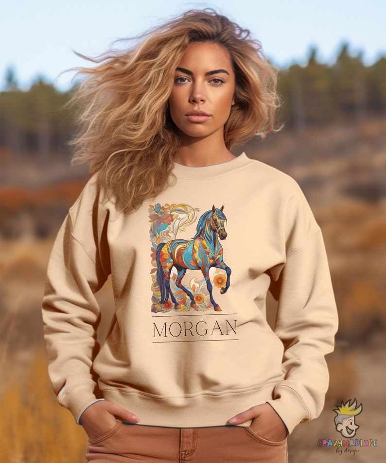 以下が含まれることがあります： ベージュのトレーナーに、カラフルな馬のグラフィックと、その下に「MORGAN」という名前がプリントされています。