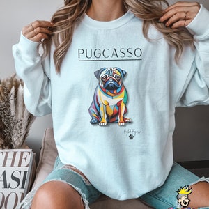 Puede incluir: Una sudadera de color azul claro con el texto "PUGCASSO" impreso en negro. Una ilustraci&oacute;n abstracta y colorida de un perro carlino est&aacute; centrada en la sudadera. El texto "Rightful Pugzasso" est&aacute; impreso debajo del perro.