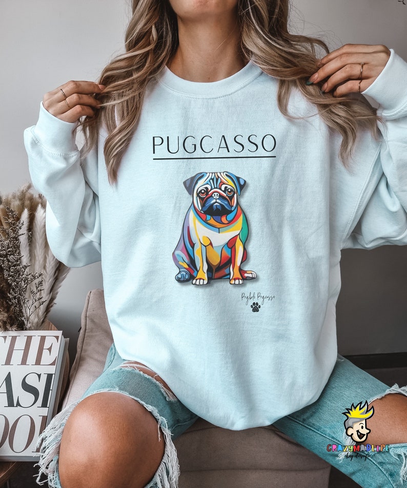 Puede incluir: Una sudadera de color azul claro con el texto "PUGCASSO" impreso en negro. Una ilustraci&oacute;n abstracta y colorida de un perro carlino est&aacute; centrada en la sudadera. El texto "Rightful Pugzasso" est&aacute; impreso debajo del perro.