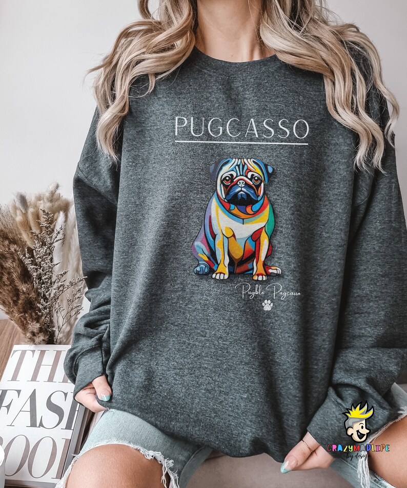 Puede incluir: Una sudadera gris oscuro con una ilustraci&oacute;n abstracta de un pug colorido y el texto "PUGCASSO" en blanco. El texto "Pugzilla Pugcasso" tambi&eacute;n est&aacute; impreso debajo de la ilustraci&oacute;n.