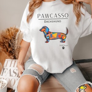 May include: White t-shirt with a colorful abstract dachshund illustration and the text "PAWCASSO" and "DACHSHUND". The text "Pawcasso" is below the dachshund.