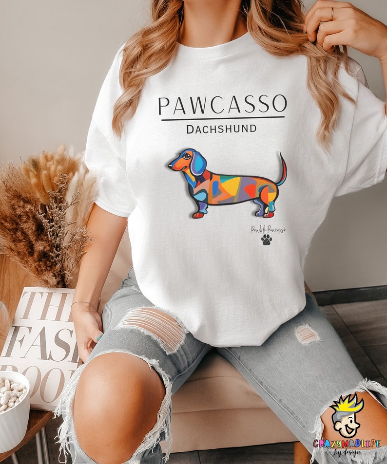 May include: White t-shirt with a colorful abstract dachshund illustration and the text "PAWCASSO" and "DACHSHUND". The text "Pawcasso" is below the dachshund.