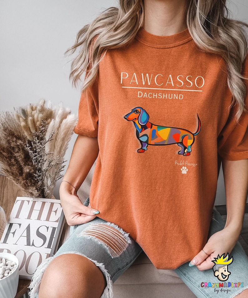 May include: A burnt orange t-shirt with the text "PAWCASSO" and "DACHSHUND" in white. A colorful abstract dachshund illustration is printed on the shirt.