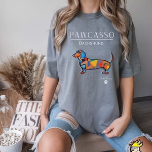 May include: Gray t-shirt with a colorful abstract dachshund illustration and the text "PAWCASSO" and "DACHSHUND". The text "Paw-casso" is below the dog and the text "DACHSHUND" is above the dog.