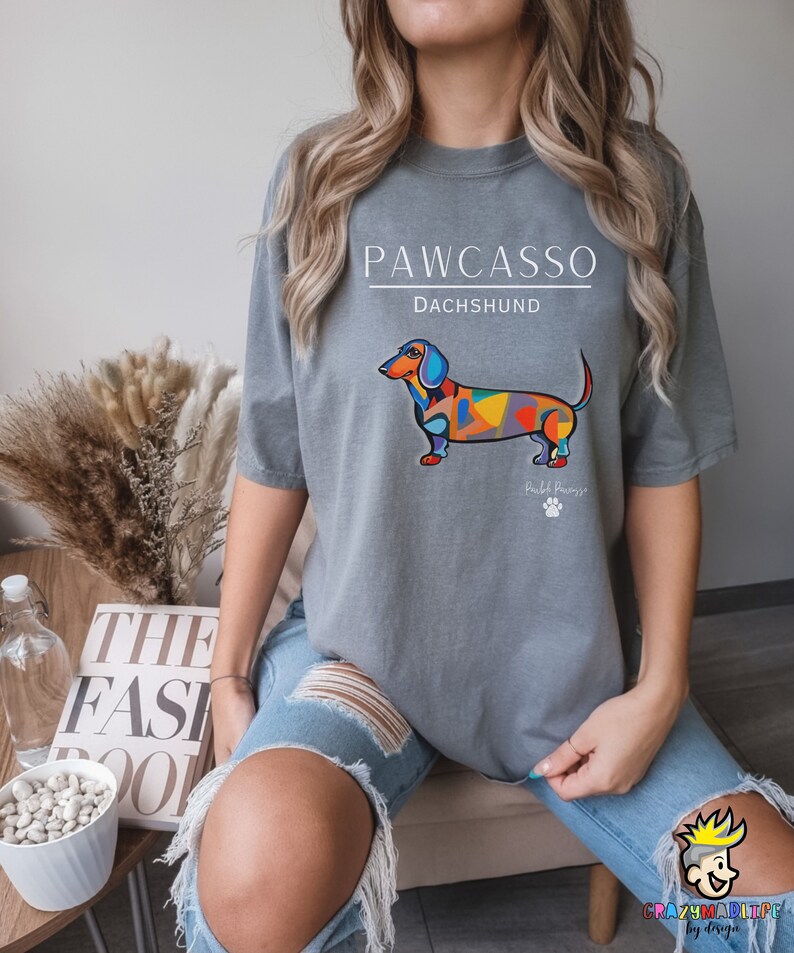May include: Gray t-shirt with a colorful abstract dachshund illustration and the text "PAWCASSO" and "DACHSHUND". The text "Paw-casso" is below the dog and the text "DACHSHUND" is above the dog.