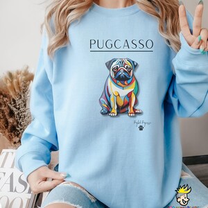 Puede incluir: Sudadera azul claro con una ilustraci&oacute;n abstracta de un carlino de colores y el texto "PUGCASSO" y "Pug-casso" en negro.