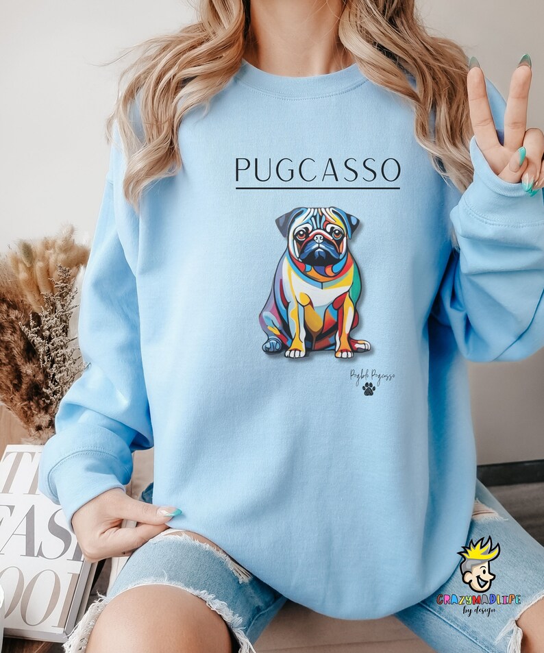 Puede incluir: Sudadera azul claro con una ilustraci&oacute;n abstracta de un carlino de colores y el texto "PUGCASSO" y "Pug-casso" en negro.