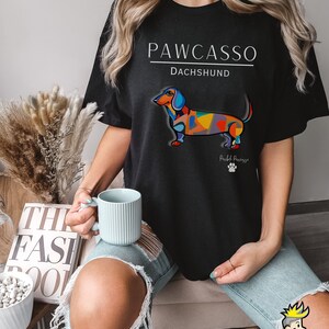 May include: Black t-shirt with a white graphic of a colorful dachshund with the text "PAWCASSO" and "DACHSHUND" above the dog and "Pawcasso" below the dog.