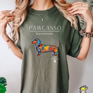 May include: Green t-shirt with a colorful dachshund illustration and the text "PAWCASSO Dachshund" printed on the front. The dachshund is in a colorful abstract style with the text "Pawcasso" below it.