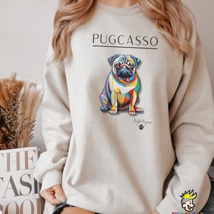 Puede incluir: Una sudadera gris claro con una ilustraci&oacute;n colorida de un perro pug. El texto "PUGCASSO" est&aacute; impreso en negro sobre el perro y "Puglife Pugasso" est&aacute; impreso en negro debajo del perro.