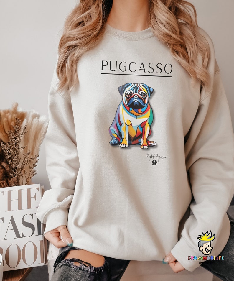 Puede incluir: Una sudadera gris claro con una ilustraci&oacute;n colorida de un perro pug. El texto "PUGCASSO" est&aacute; impreso en negro sobre el perro y "Puglife Pugasso" est&aacute; impreso en negro debajo del perro.