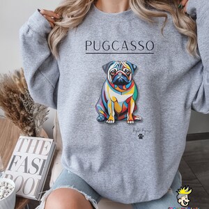 Puede incluir: Sudadera gris con un gr&aacute;fico colorido de un pug con el texto "PUGCASSO" y el texto "By Puggy" debajo del gr&aacute;fico.