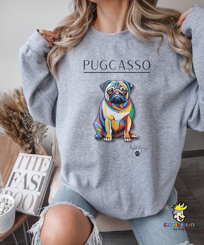 Puede incluir: Sudadera gris con un gr&aacute;fico colorido de un pug con el texto "PUGCASSO" y el texto "By Puggy" debajo del gr&aacute;fico.