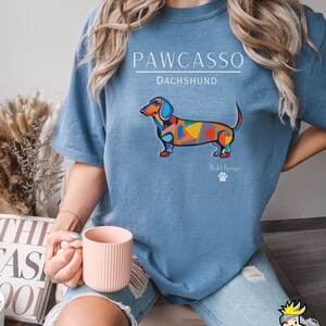 May include: A blue t-shirt with a colorful dachshund illustration and the text "PAWCASSO" and "DACHSHUND". The text "Pawcasso" is written in a smaller font below the dachshund.