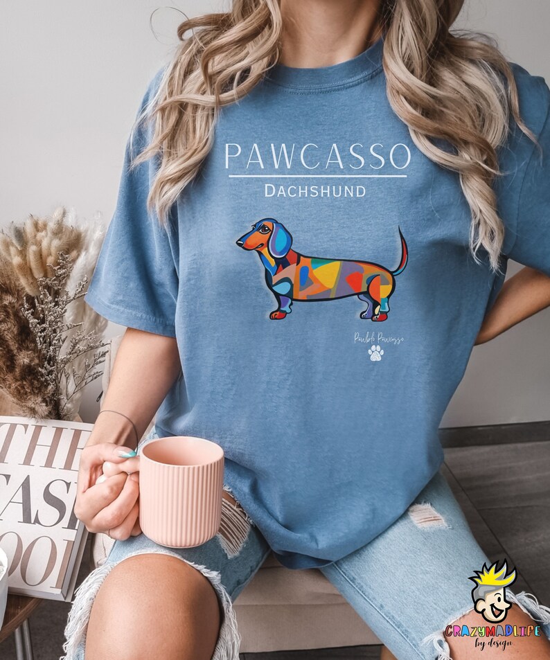 May include: A blue t-shirt with a colorful dachshund illustration and the text "PAWCASSO" and "DACHSHUND". The text "Pawcasso" is written in a smaller font below the dachshund.