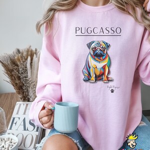Puede incluir: Una sudadera rosa con una ilustraci&oacute;n abstracta de un pug colorido y el texto "PUGCASSO" impreso en la parte delantera. El texto "Pugb Picasso" est&aacute; impreso debajo de la ilustraci&oacute;n.