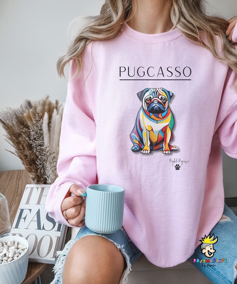 Puede incluir: Una sudadera rosa con una ilustraci&oacute;n abstracta de un pug colorido y el texto "PUGCASSO" impreso en la parte delantera. El texto "Pugb Picasso" est&aacute; impreso debajo de la ilustraci&oacute;n.