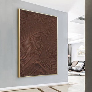 Puede incluir: Una pintura abstracta marr&oacute;n con una superficie texturizada, enmarcada en un marco dorado. La pintura est&aacute; colgada en una pared blanca.