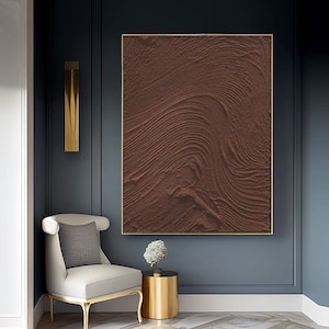 Puede incluir: Una pintura abstracta marr&oacute;n con una superficie texturizada y l&iacute;neas en espiral. La pintura est&aacute; enmarcada en un marco dorado y cuelga de una pared azul oscuro.