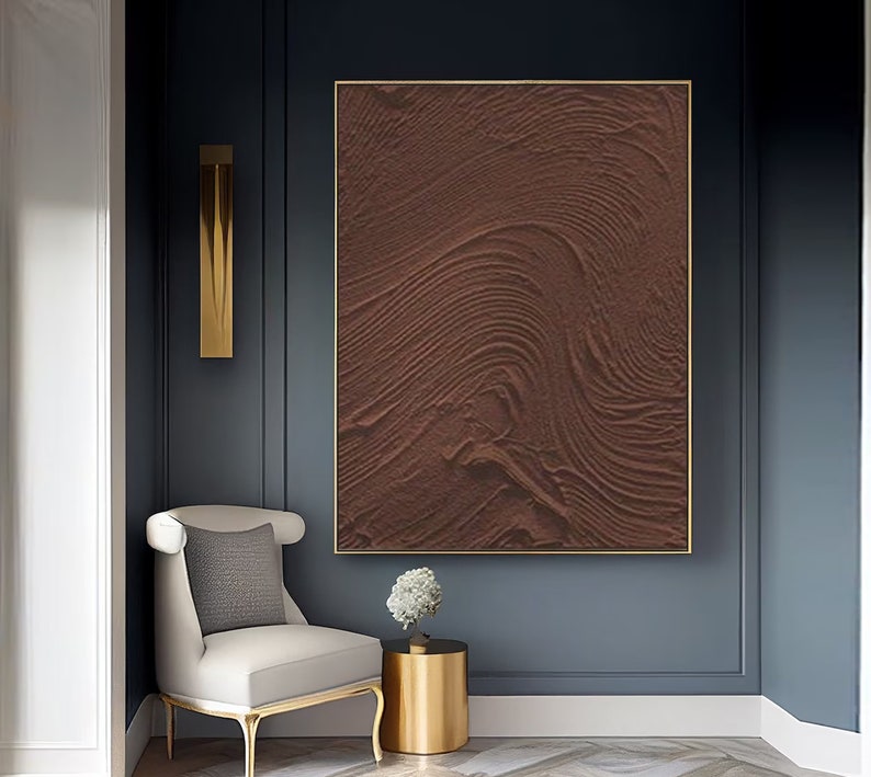Puede incluir: Una pintura abstracta marr&oacute;n con una superficie texturizada y l&iacute;neas en espiral. La pintura est&aacute; enmarcada en un marco dorado y cuelga de una pared azul oscuro.