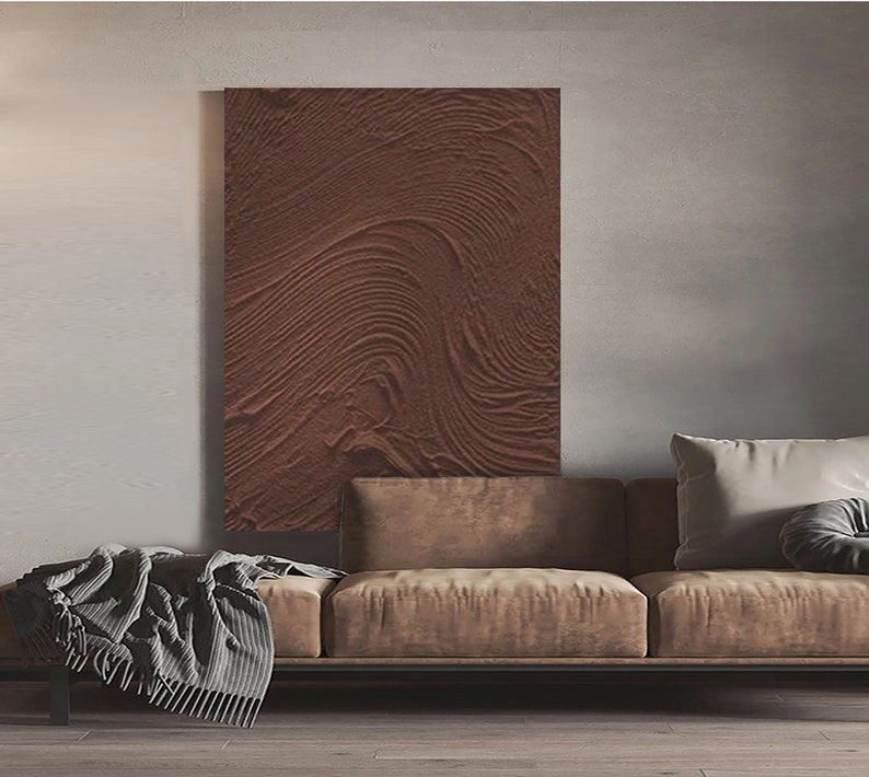 Puede incluir: Un sof&aacute; de cuero marr&oacute;n con dos almohadas, una blanca y otra gris, frente a una pieza de arte de pared de textura marr&oacute;n.