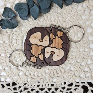 Könnte beinhalten: Zwei Holzschlüsselanhänger mit Pinguin-Design. Die Schlüsselanhänger sind aus dunkelbraunem Holz mit einer helleren Holzeinlage für den Körper des Pinguins gefertigt. Jeder Schlüsselanhänger hat einen silberfarbenen Metallring und eine Kette.
