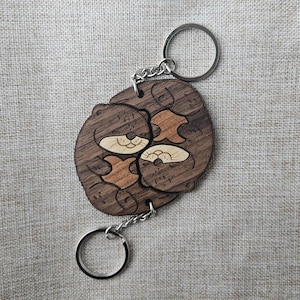 Könnte beinhalten: Ein Holzschlüsselanhänger mit einem Design von zwei Ottern. Die Otter sind hellbraun mit cremefarbenen Gesichtern und vor einem dunkelbraunen Hintergrund. Der Schlüsselanhänger hat zwei silberne Schlüsselringe und eine silberne Kette.