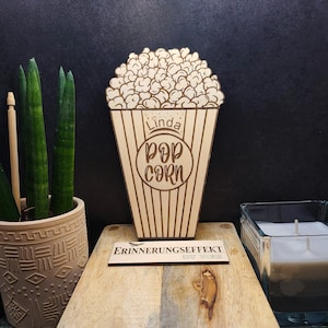 Op de afbeelding: Een houten popcornbox met de naam "Linda" en de woorden "Pop Corn" erop gegraveerd. De doos is versierd met een hartje en sterren. De doos staat op een houten oppervlak met een klein bordje waarop "Erinnerungseffekt by Tobi" staat.