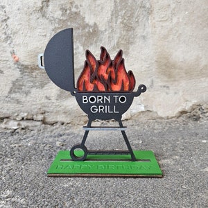 K&ouml;nnte beinhalten: Ein schwarzer und roter Grill aus Holz mit Flammen und dem Text "Born to Grill" auf der Vorderseite. Der Grill steht auf einem gr&uuml;nen Sockel mit dem Text "Happy Birthday".