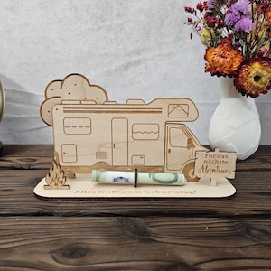 May include: A wooden campervan-shaped money holder with a cloud and flame design. The holder has the German text "Alles Gute zum Geburtstag!" and "F&uuml;r das n&auml;chste Abenteuer". A rolled-up banknote is held in place.