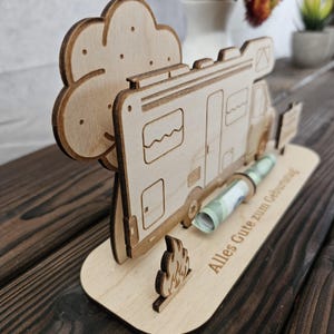 May include: A wooden money holder shaped like a camper van, with a tree and campfire cutouts. The base is engraved with the words "Alles Gute zum Geburtstag". A rolled-up banknote is in the slot.