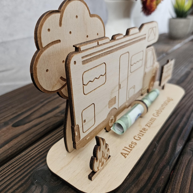 May include: A wooden money holder shaped like a camper van, with a tree and campfire cutouts. The base is engraved with the words "Alles Gute zum Geburtstag". A rolled-up banknote is in the slot.