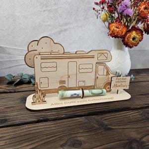May include: A wooden money holder shaped like a camper van, with a rolled-up banknote held in place. The design includes a campfire, a cloud, and German text: "Alles Gute zum Geburtstag!" and "F&uuml;r das n&auml;chste Abenteuer".