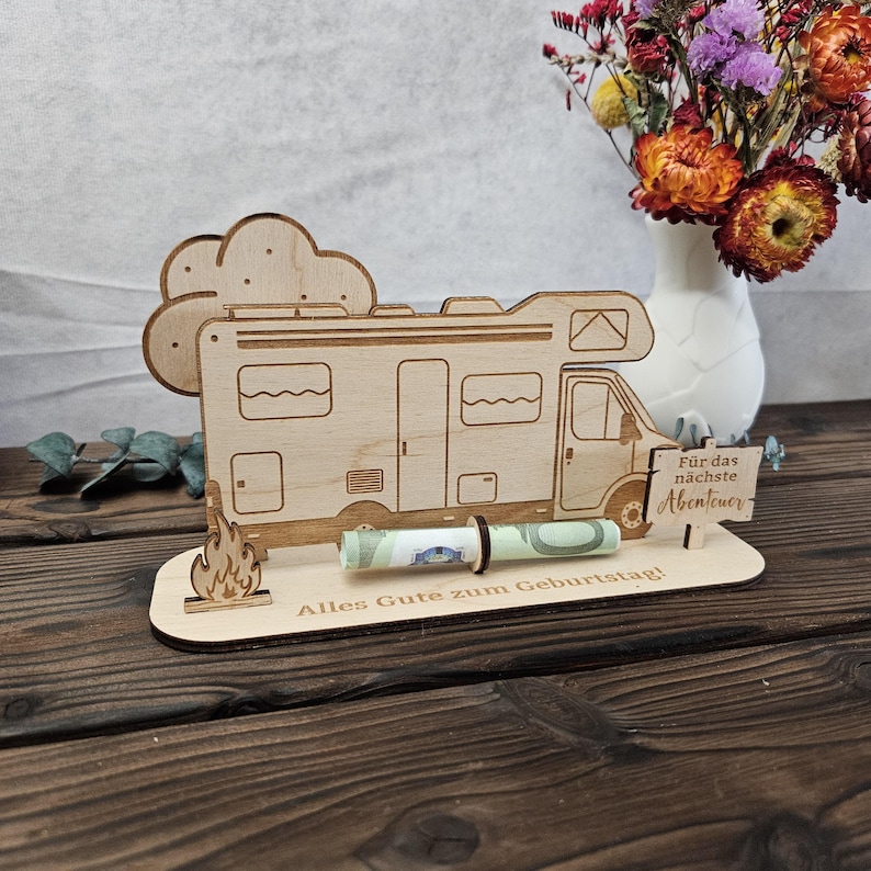 May include: A wooden money holder shaped like a camper van, with a rolled-up banknote held in place. The design includes a campfire, a cloud, and German text: "Alles Gute zum Geburtstag!" and "F&uuml;r das n&auml;chste Abenteuer".